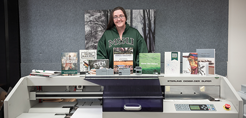 Woman standing in MSU sweat shirt behind a book publishing machine